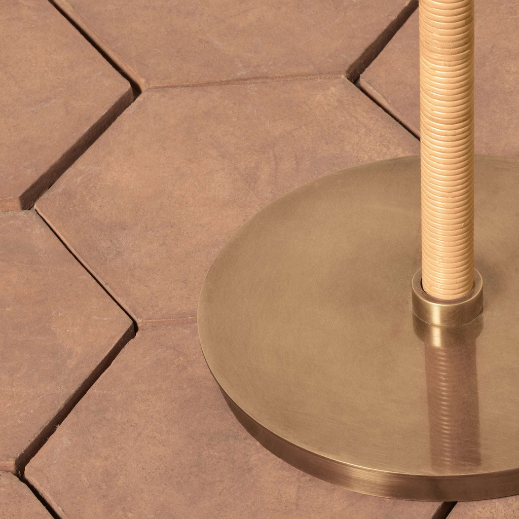Close-up of a brass Delphine floor lamp base with a ribbed pole on a terracotta hexagonal tile floor.