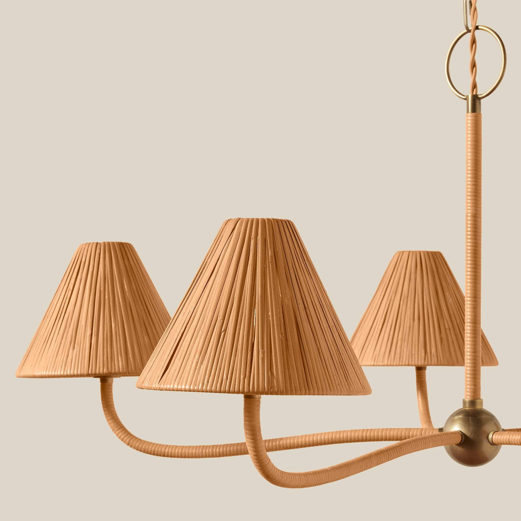 A close-up of a modern Delphine chandelier with three conical woven rattan shades and curved brass metal arms, set against a solid light beige background.