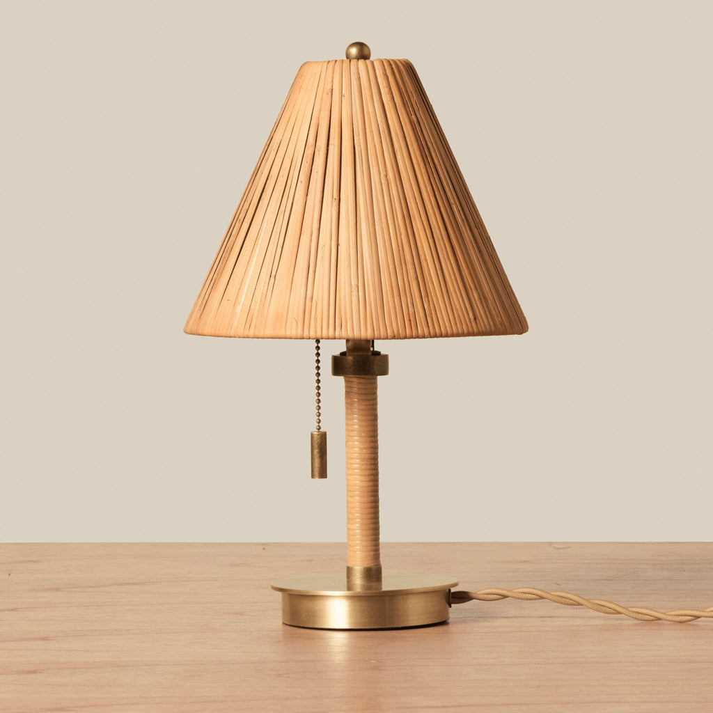 A vintage-style Delphine Mini table lamp with a pleated beige fabric shade, a wrapped beige stem, and a brass base, shown on a wooden surface against a neutral beige background.
