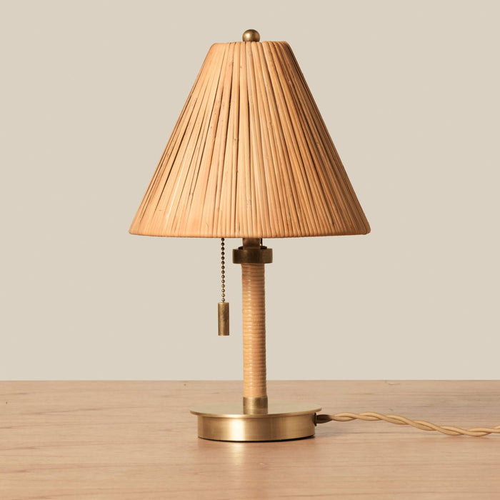 A vintage-style Delphine Mini table lamp with a pleated beige fabric shade, a wrapped beige stem, and a brass base, shown on a wooden surface against a neutral beige background.