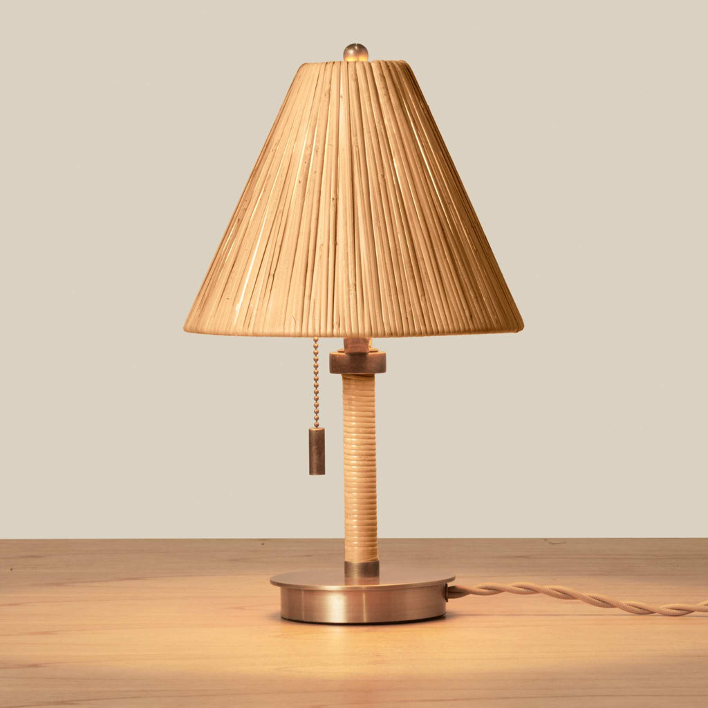 A Delphine Mini table lamp with a pleated beige fabric shade, a wrapped natural wood stem, and a round metallic base, shown on a wooden tabletop against a neutral beige background.