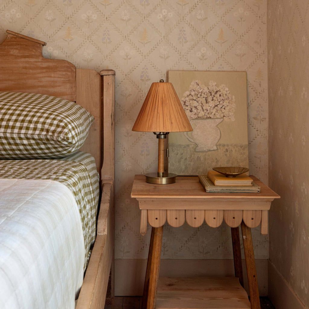 Wooden bedside table with Mini Delphine table lamp and books next to a bed with checkered pillow and green bedding.
