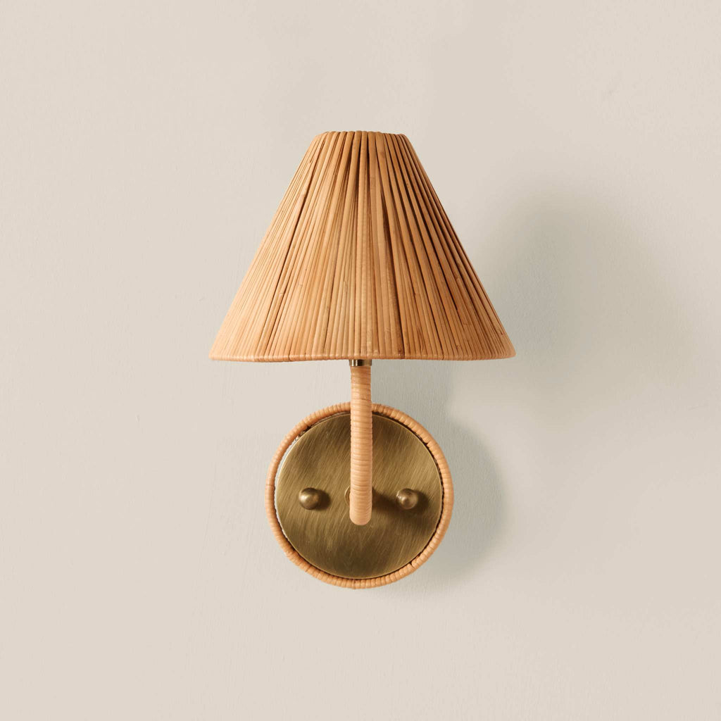 A wall-mounted Delphine Sconce light fixture with a pleated natural bamboo shade, a wrapped bamboo stem, and a round brass wall plate, shown against a solid light beige background.