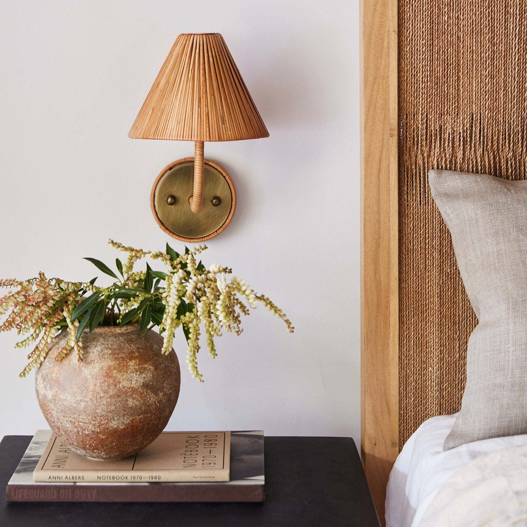 Decorative Delphine Sconce with woven shade on a wall next to a vase with flowers and books on a table.