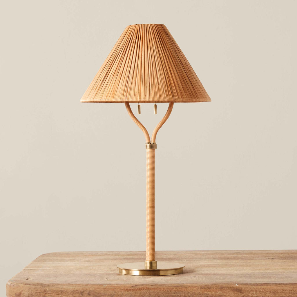 A Delphine table lamp with a pleated bamboo shade, a curved bamboo stem, and a brass base, shown on a wooden surface against a neutral beige background.