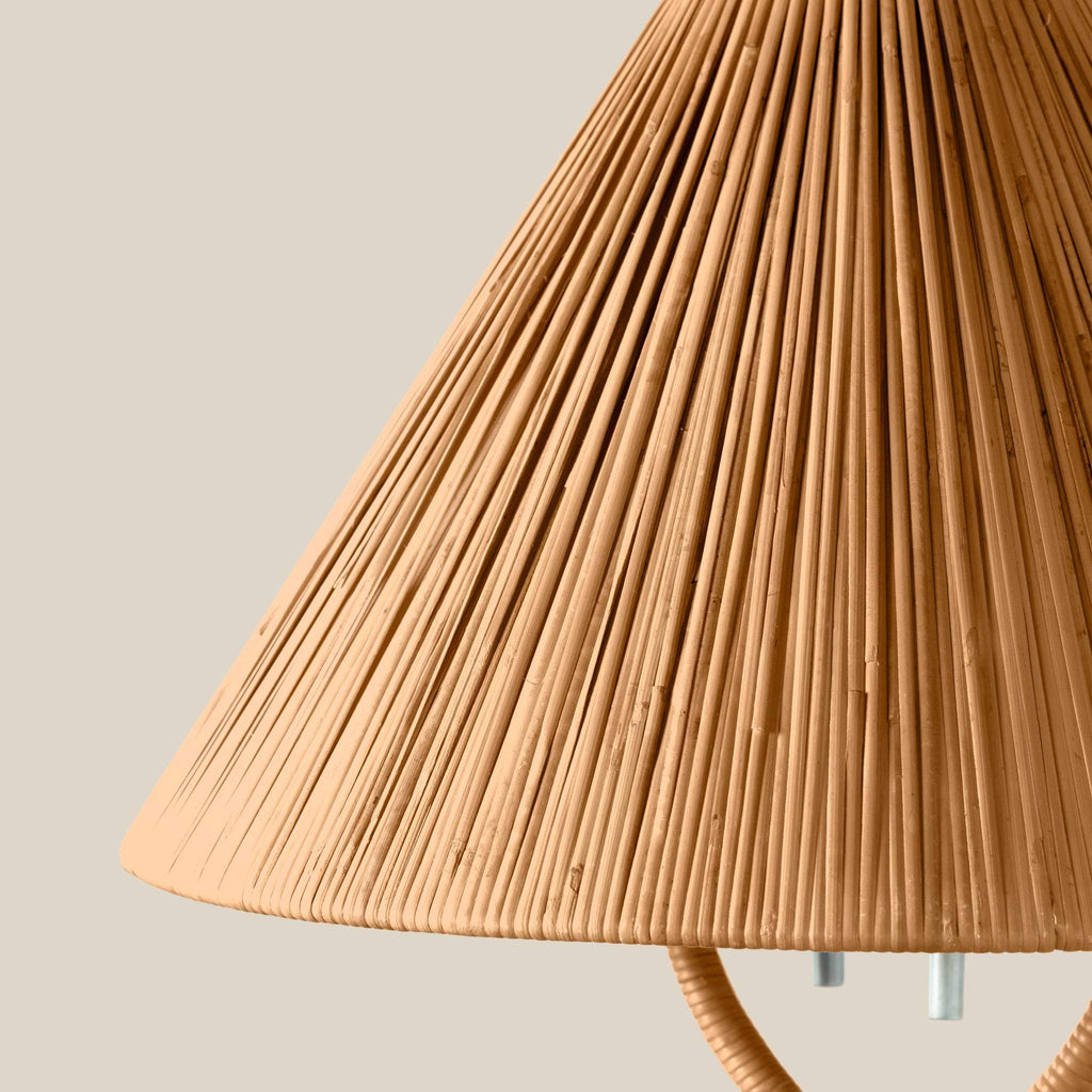 Close-up of a pleated conical Delphine table lampshade in a warm natural tan color, with part of the curved lamp base visible against a plain light beige background.