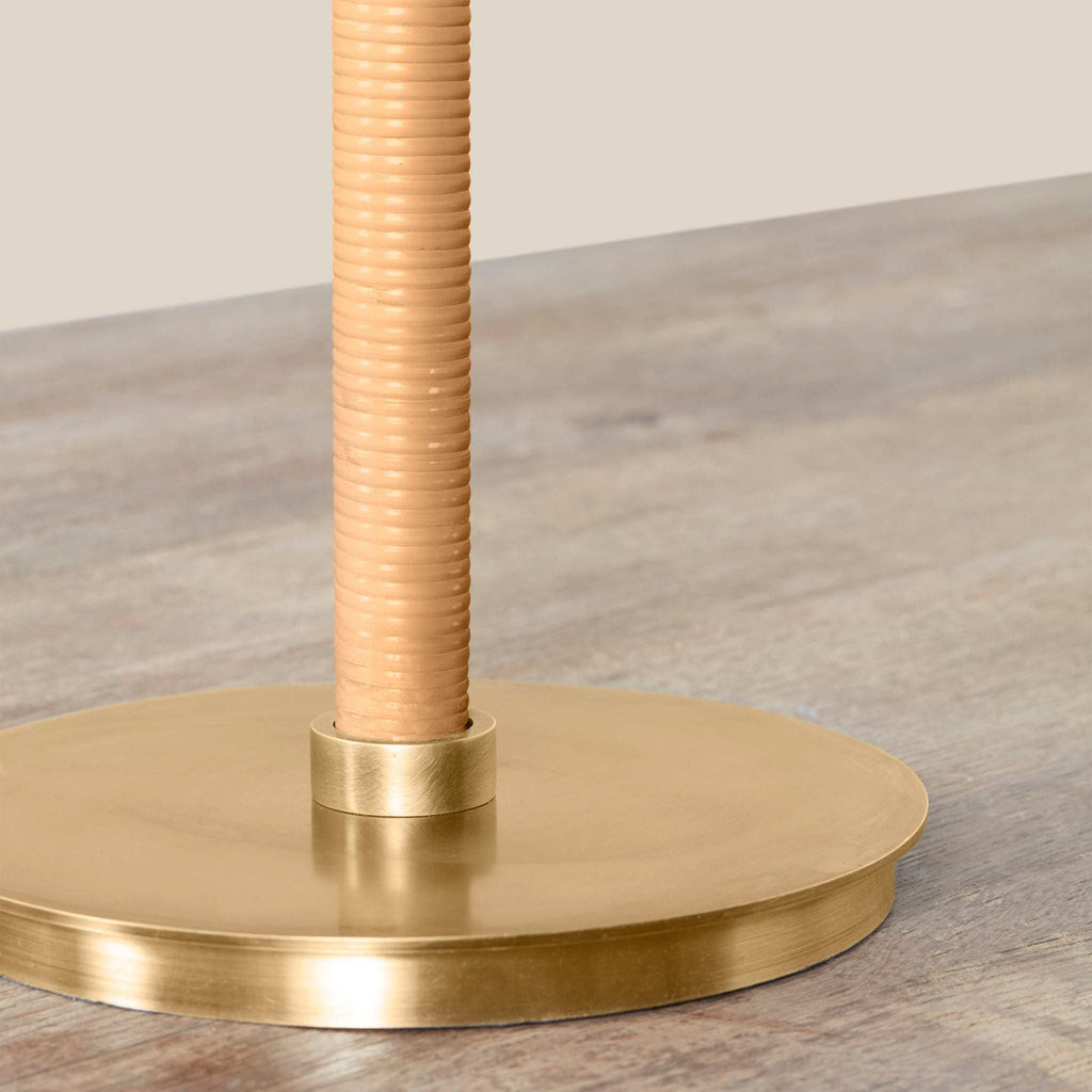 Close-up of a Delphine table lamp base with a round brushed brass platform and a ribbed beige fabric-covered pole, shown on a wood surface against a neutral beige background.