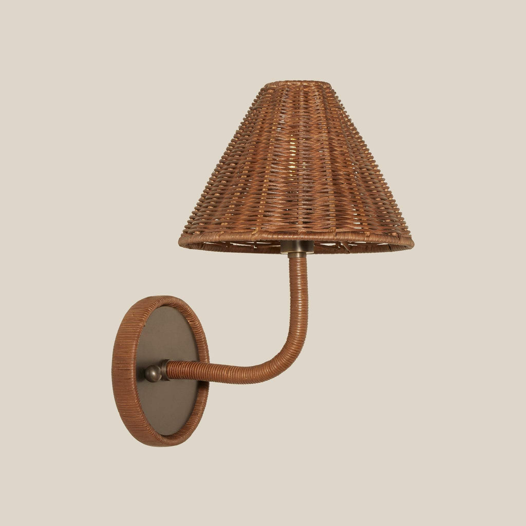 Delphine wicker wall sconce with a conical woven shade and a curved arm mounted on a round pewter backplate, shown against a solid beige background.