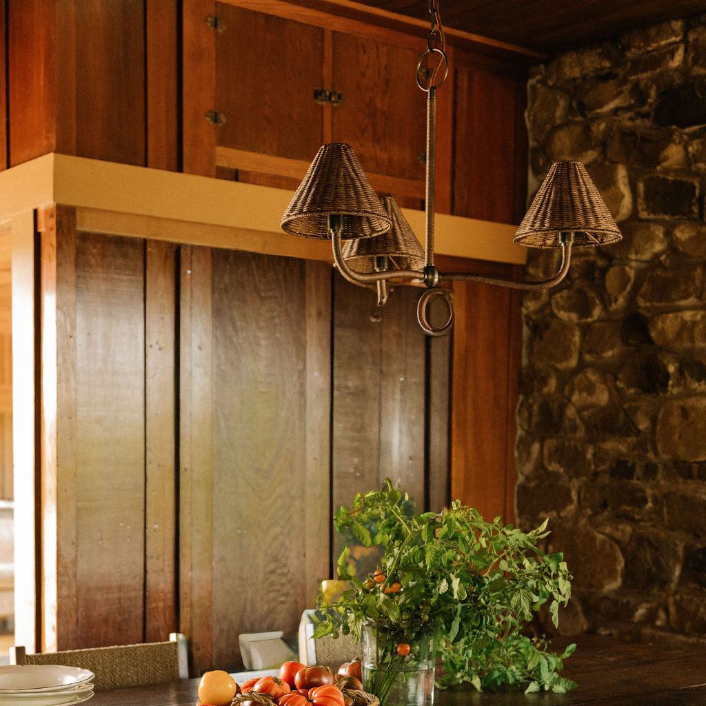 Delphine three-light pendant chandelier with woven wicker shades and a pewter metal frame hangs in a warm, wood-paneled kitchen with a stone wall and a table with tomatoes and herbs below.