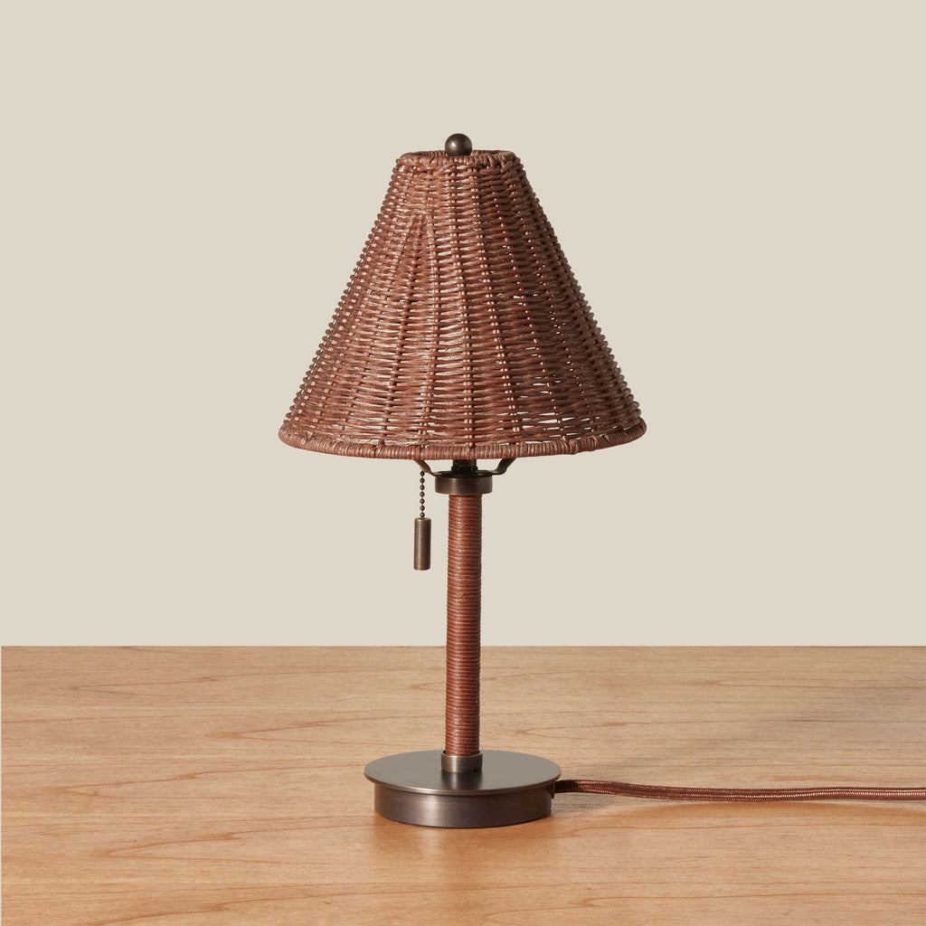 A small Delphine wicker table lamp with a brown woven wicker shade, a matching brown wrapped stem, and a round dark metal base, shown on a wooden surface against a plain beige background.