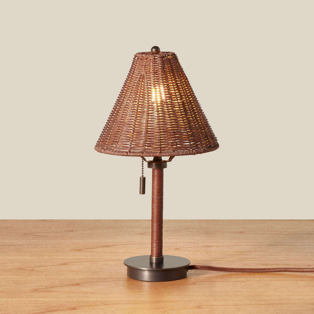 A Delphine table lamp with a woven brown wicker shade, a wrapped brown stem, and a round dark metal base, shown lit on a wooden surface against a plain beige background.