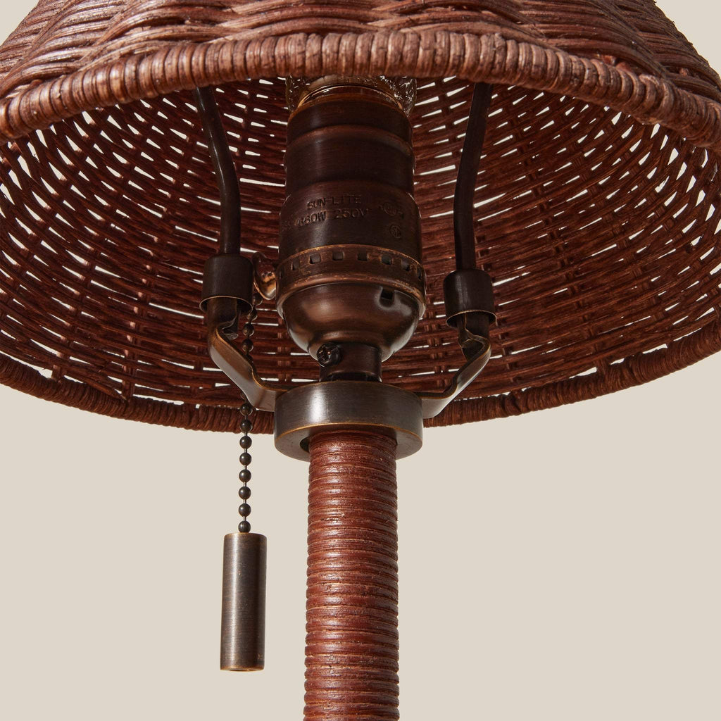 Close-up of a Delphine table lamp with a woven wicker shade, a wrapped brown stem, and a brass pull chain against a plain beige background.
