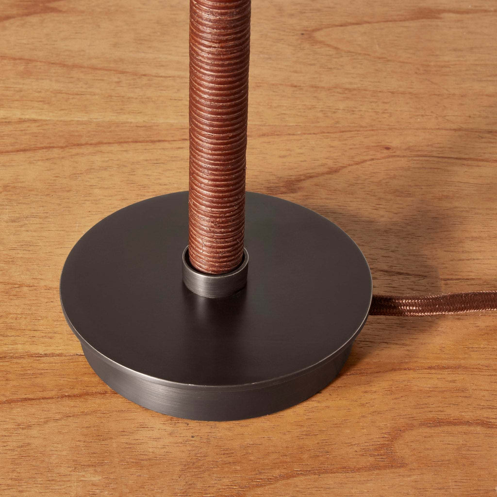 A close-up of a Delphine table lamp base with a round matte black metal base and a textured copper-colored vertical stem, shown on a wooden tabletop.