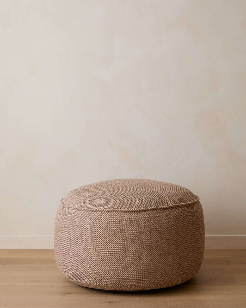 A round, low-profile pouf ottoman upholstered in a textured beige woven fabric, set against a minimalist beige wall and light wood floor.