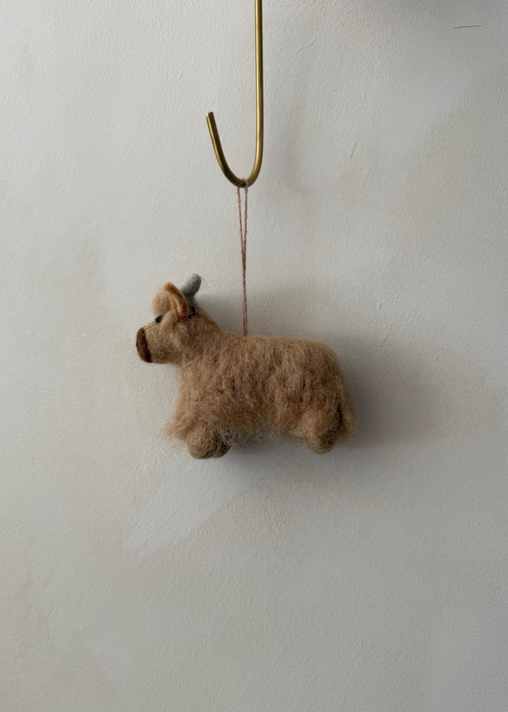 Small brown felt shetland cow ornament hanging on a lime washed wall. Saffron + Poe