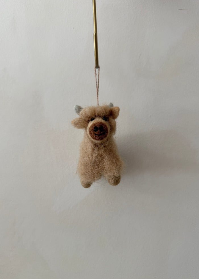 Small brown felt shetland cow ornament hanging in front of a lime washed wall. Saffron + Poe