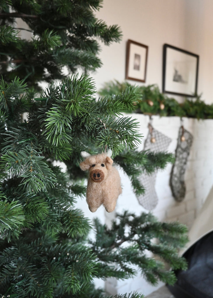 Small brown felt shetland cow ornament hanging on a Christmas Tree facing the camera. Saffron + Poe