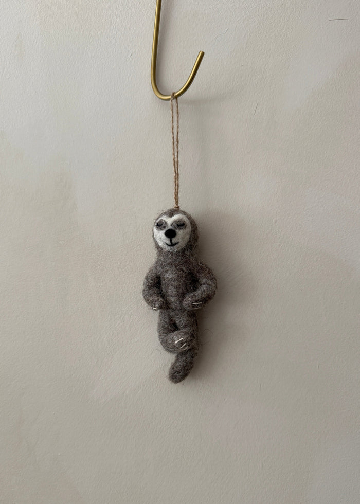 Felted yoga sloth ornament hanging from a hook on a lime washed wall. Saffron + Poe