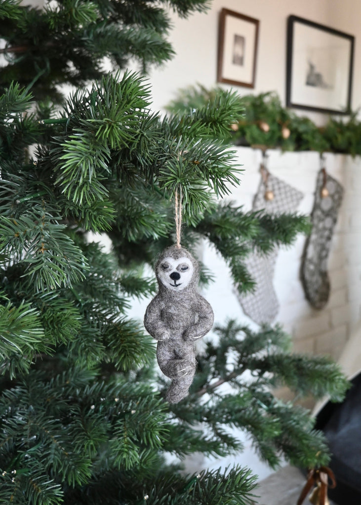 Felted yoga sloth ornament hanging on a Christmas tree with stockings in the background. Saffron + Poe