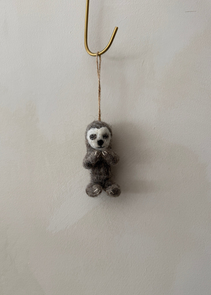 Felted yoga sloth ornament hanging on a hook against a lime washed wall. Saffron + Poe