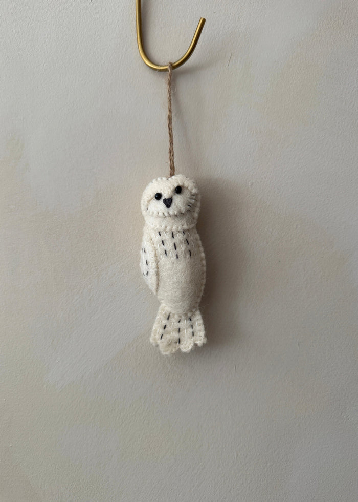 Knitted owl felt ornament hanging on a hook against a lime washed wall. Saffron + Poe