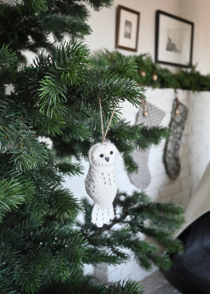 White owl felt ornament hanging on a Christmas tree with a fireplace in the background. Saffron + Poe