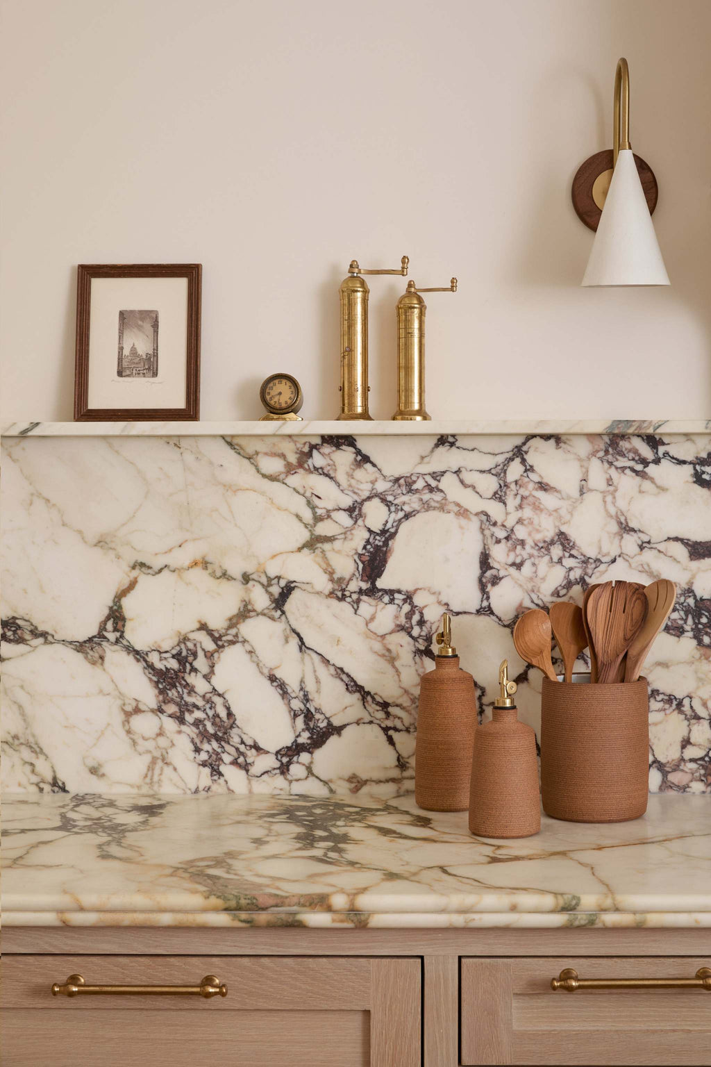 Terracotta-colored ceramic utensil jar with wooden utensils and olive oil and vinegar pourer arranged on the counter in a styled kitchen counter scene with a white and gray veined marble backsplash and matching marble countertop. A white wall sconce, framed art, and salt and pepper grinders. 