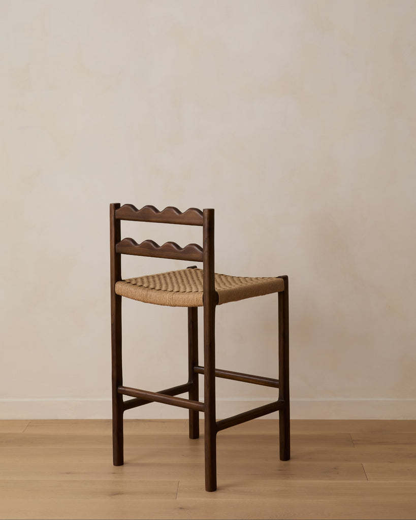 Ionian scalloped counter stool with woven seat on white oak flooring against a beige wall. Saffron and Poe