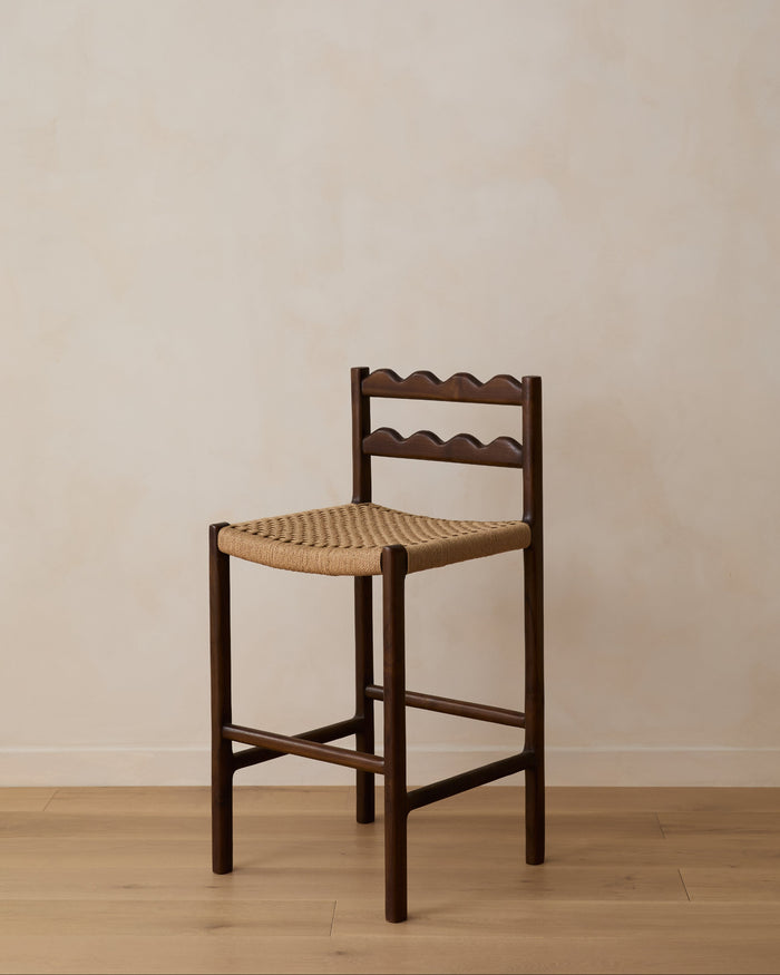 Ionian scalloped counter stool with woven seat on white oak flooring against a beige wall. Saffron and Poe