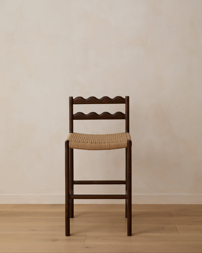 Ionian scalloped counter stool with woven seat on white oak flooring against a beige wall. Saffron and Poe