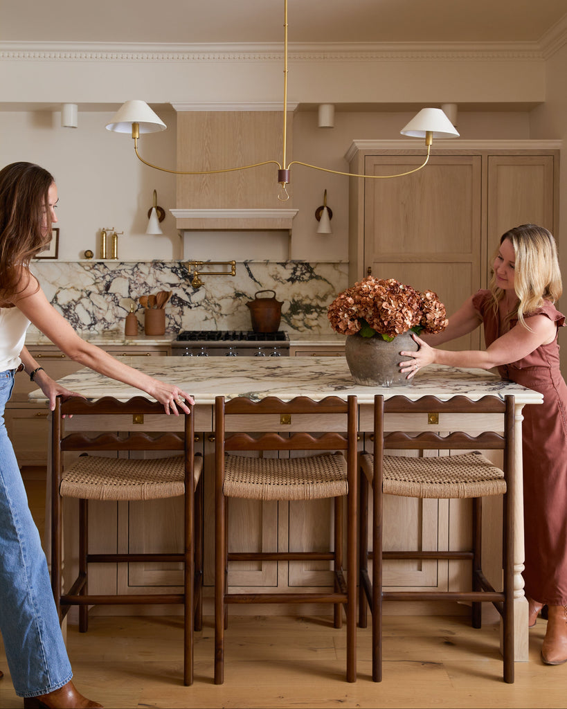 Ionian scalloped counter stool with woven seat in a white oak kitchen with marble counters and two people styling. Saffron and Poe