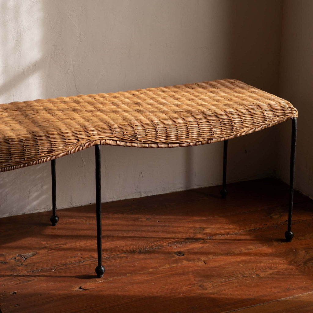 Lacoste woven rattan bench with black metal iron legs on a wooden floor against a white plaster wall