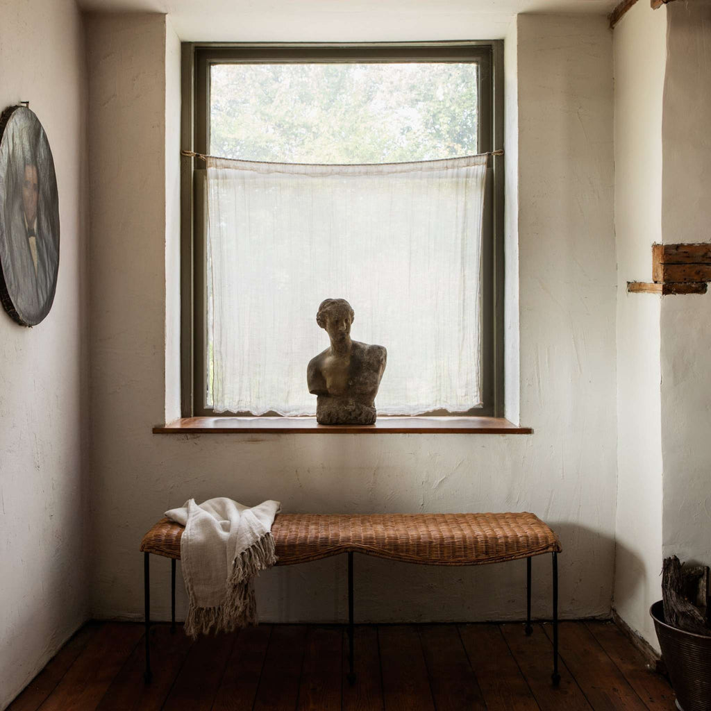 Lacoste woven rattan bench with a white blanket in a European-style living room with a bust statue on windowsill