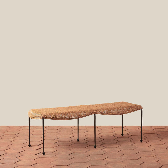 Woven rattan Lacoste bench with wrought iron legs on a terracotta tile floor with a beige wall background.