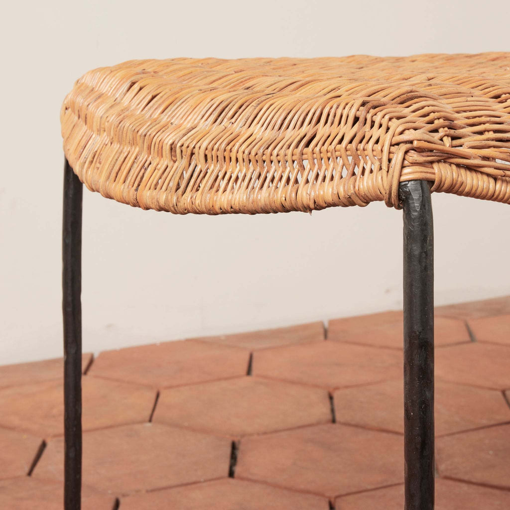 Close up detail of Lacoste indoor outdoor woven rattan bench with black iron legs against terracotta tile floor and white background.