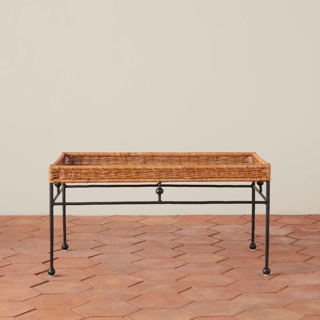Lacoste rectangular wicker tray table with a black iron frame and legs, set against a neutral beige wall and terracotta tile floor