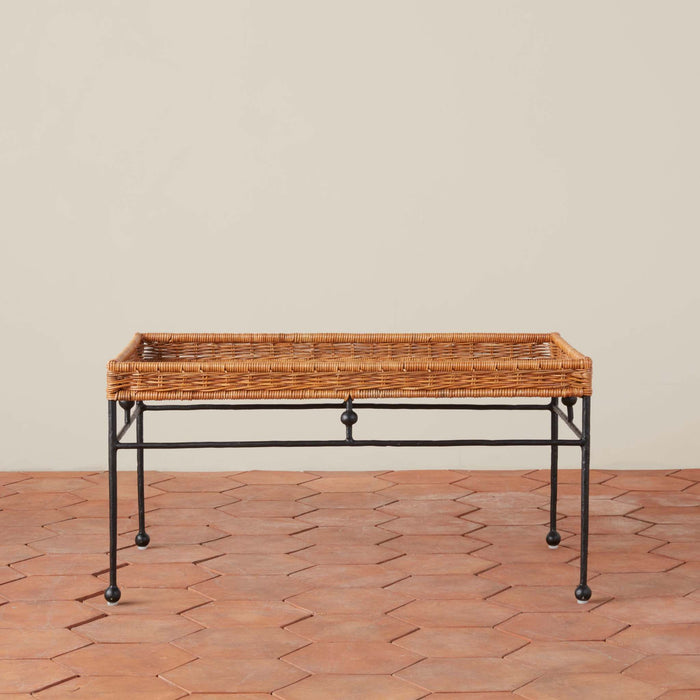Lacoste rectangular wicker tray table with a black iron frame and legs, set against a neutral beige wall and terracotta tile floor
