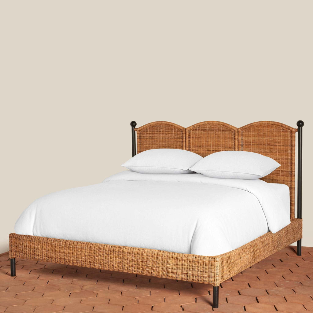 Lacoste Rattan King Bed with scallop-shaped headboard, black iron frame, and white bedding on a terracotta tile floor.