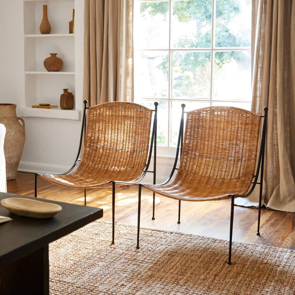 Two woven Lacoste lounge chairs with woven wicker seats and black metal frames, placed in a bright living room with wood floors, beige curtains, and a window.