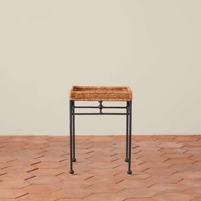 Lacoste rectangular rattan tray table with a black metal frame and legs, set against a neutral beige wall and terracotta hexagonal tile floor.