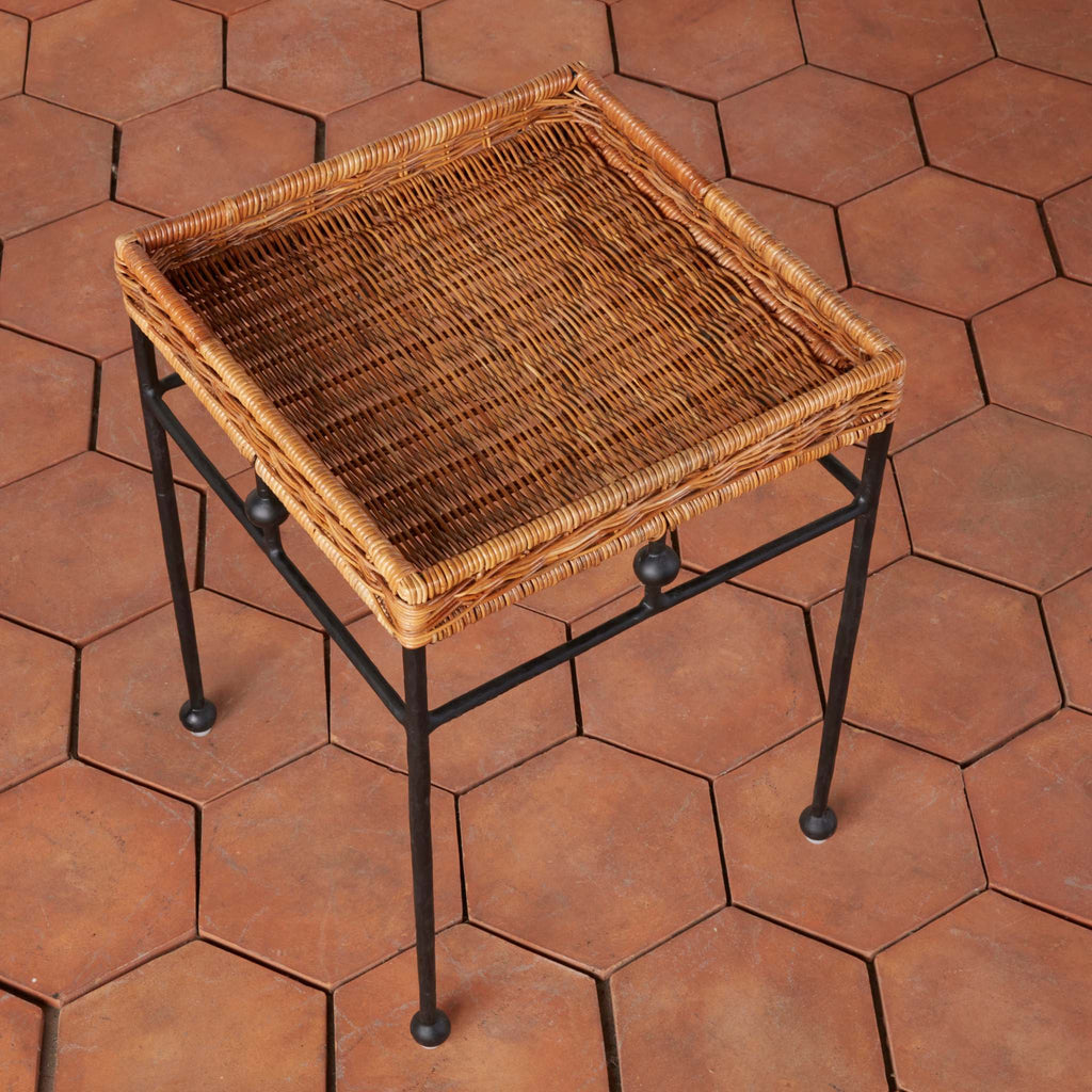 Lacoste side table with a natural brown woven rattan top and a black metal frame, shown on terracotta hexagonal tiles.