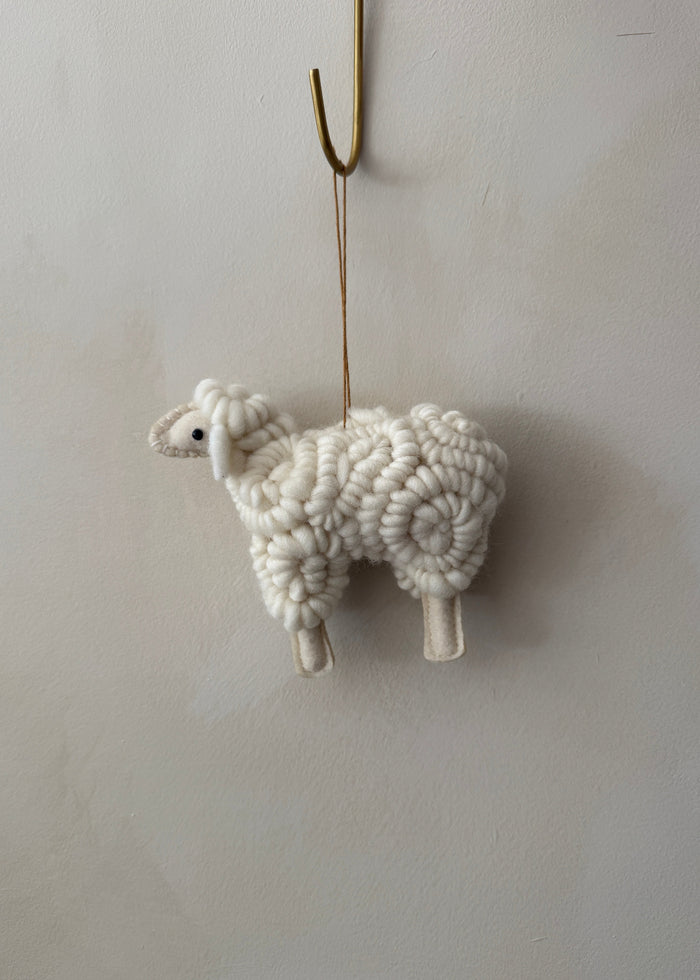 Knitted felt sheep ornament hanging on a hook against a lime washed wall. Saffron + Poe