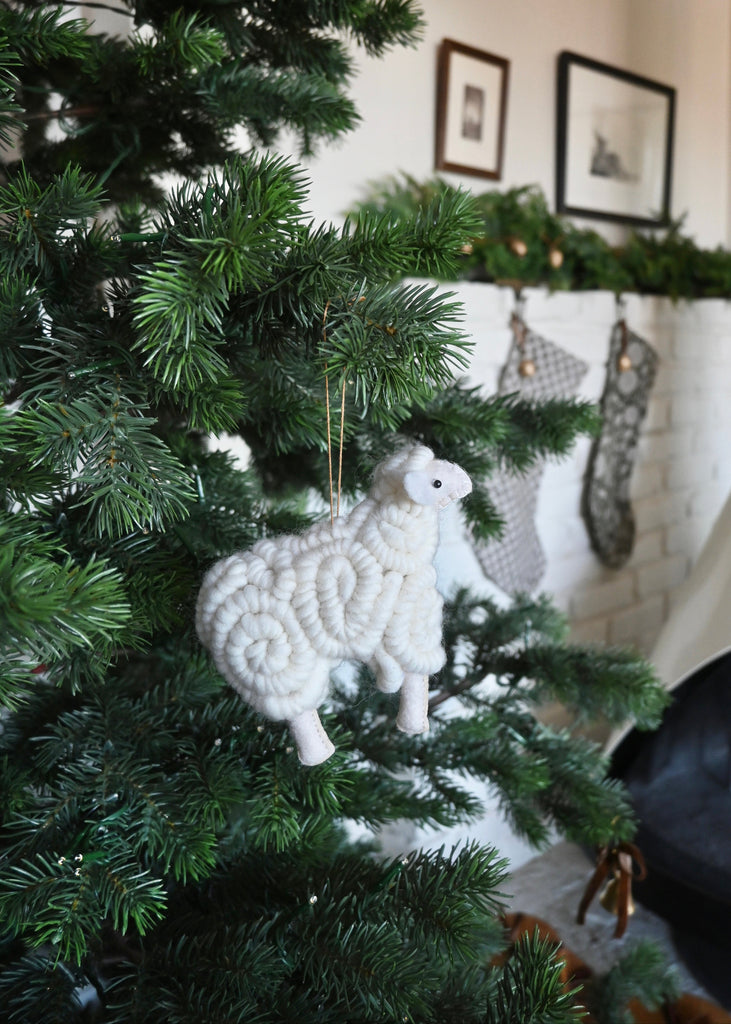 White knitted sheep ornament on a Christmas tree with a fireplace and stockings in the background. Saffron + Poe