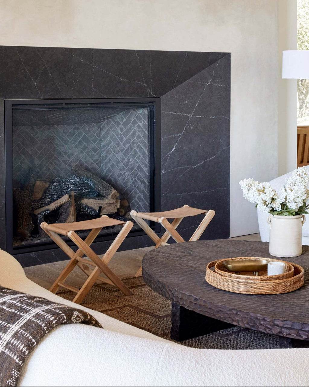 A styled interior scene featuring a dark charcoal stone fireplace with a black surround, a round dark woven coffee table, two leather folding stools, a white sofa with pillows, and a vase of white flowers on a dark coffee table.
