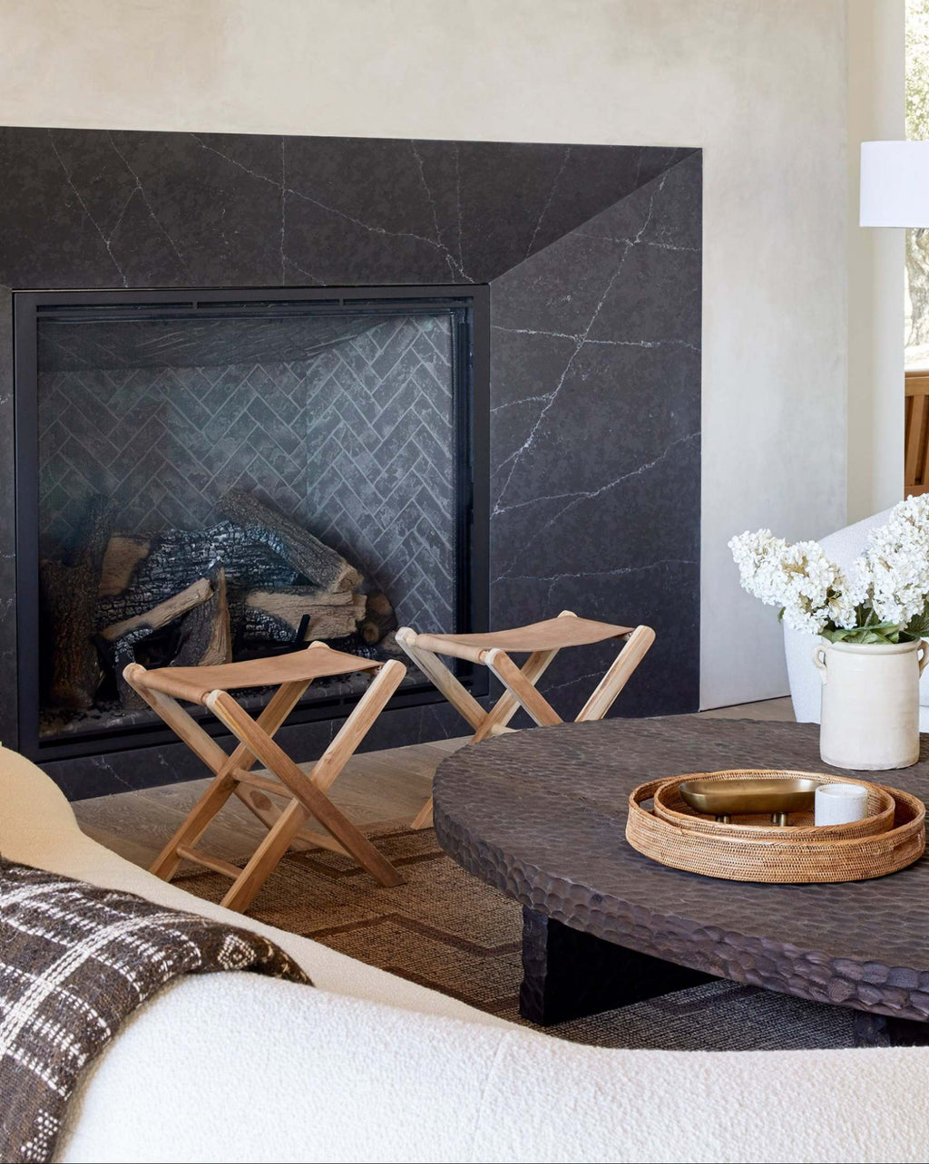 A styled interior scene featuring a dark charcoal stone fireplace with a black surround, a round dark woven coffee table, two leather folding stools, a white sofa with pillows, and a vase of white flowers on a dark coffee table.