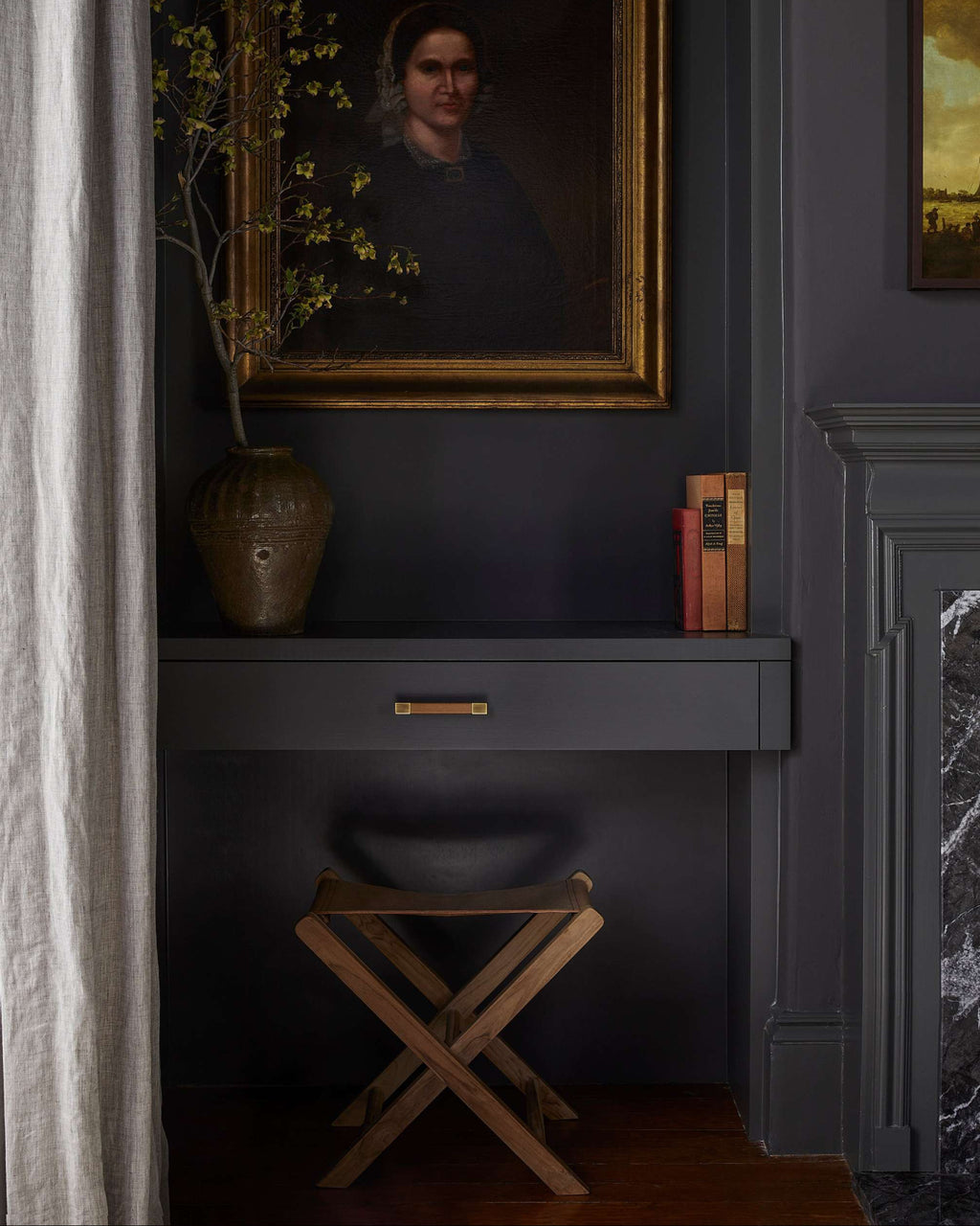 A dark charcoal gray console table with a single drawer and brass pull, styled in a moody room with a leather folding stool, a ceramic vase with yellow blossoms, and a large framed portrait on the wall above.