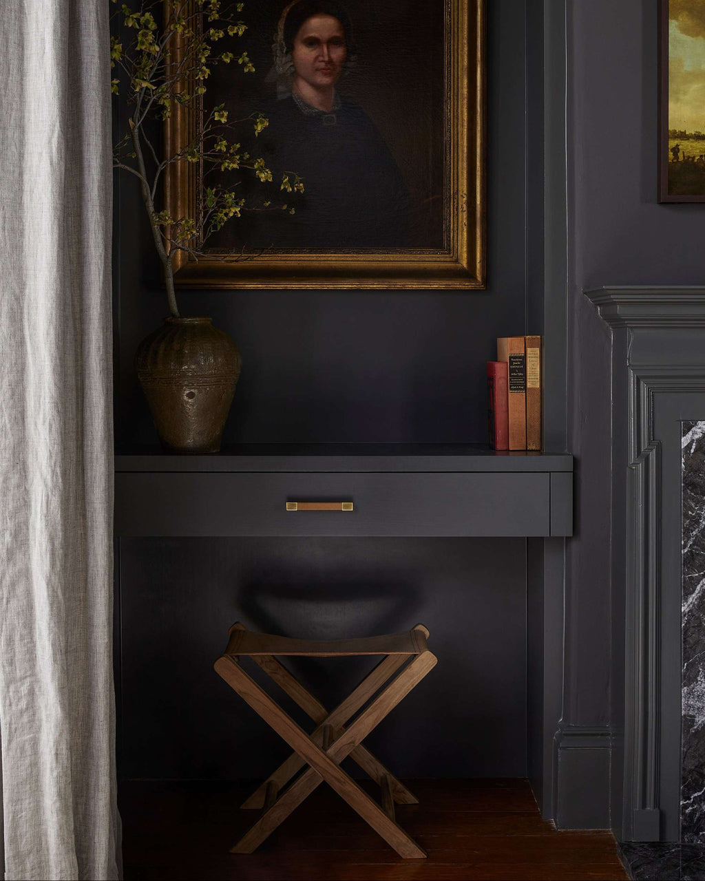 A dark charcoal gray console table with a single drawer and brass pull, styled in a moody room with a leather folding stool, a ceramic vase with yellow blossoms, and a large framed portrait on the wall above.