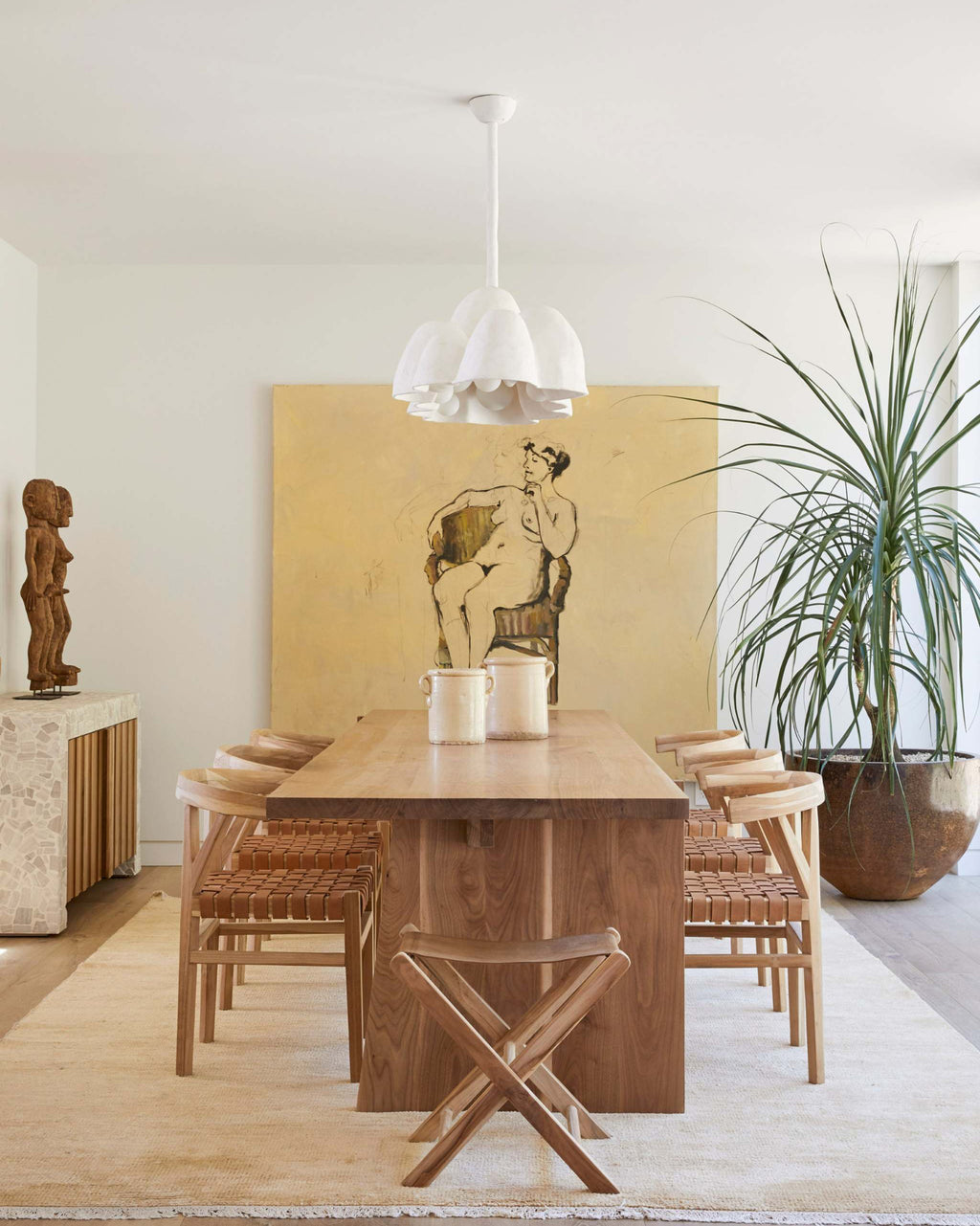 A mid-century modern dining room setup featuring a rectangular wooden dining table and four matching woven leather wishbone dining chairs and a leather folding stools, under a white multi-light pendant lamp. 