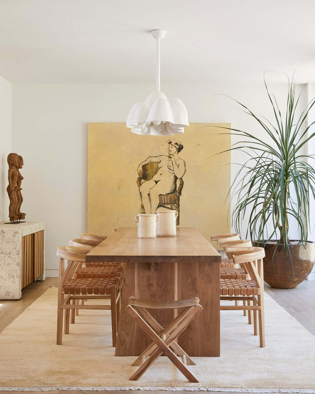 A mid-century modern dining room setup featuring a rectangular wooden dining table and four matching woven leather wishbone dining chairs and a leather folding stools, under a white multi-light pendant lamp. 