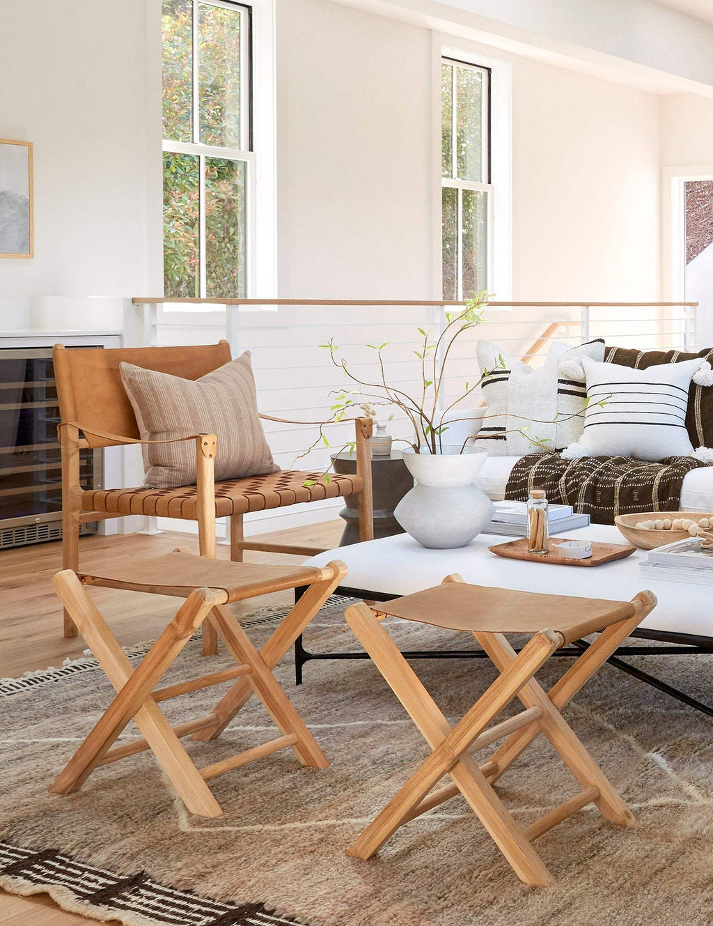 Two light natural wood folding director-style leather folding stools shown in a bright modern living room beside a white coffee table and sofa.